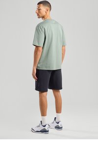 Light green cotton t-shirt with short sleeves, straight cut. Black shorts, white sneakers with blue and grey accents. Plain background.