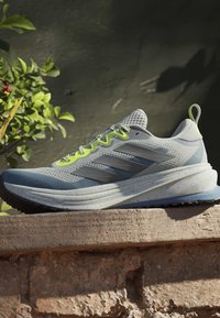 Chaussure de sport gris clair avec des rayures noires et des accents de lacets jaune néon posée sur un rebord en pierre avec un feuillage vert en arrière-plan.