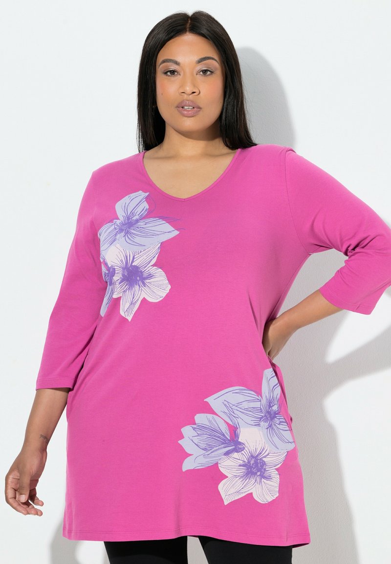 Kvinde iført en lys pink langærmet tunika med lilla og hvide blomsterprint, poser med den ene hånd på hoften mod en hvid baggrund.