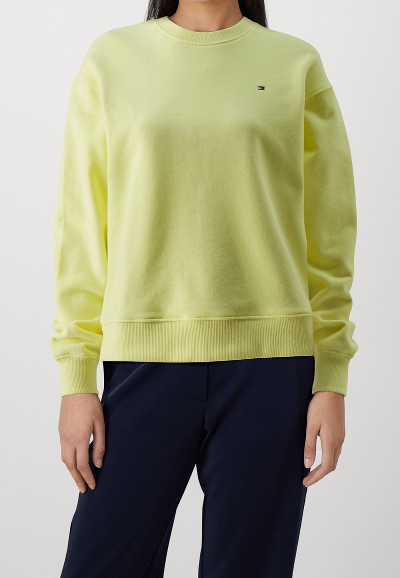 Personne portant un sweat-shirt jaune clair à manches longues et un pantalon bleu marine foncé, debout avec les bras détendus le long du corps.