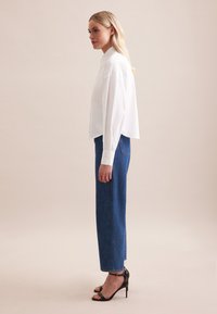 Camicetta bianca a maniche lunghe e corto, abbinata a jeans in denim blu a vita alta. Sandali neri con tacco completano l'outfit.
