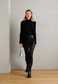 Blazer noir sur un top noir, jupe en simili cuir avec deux anneaux argentés, collants noirs, bottes noires jusqu'au genou, tenant un sac argenté.
