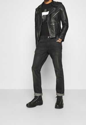 Man in een zwart leren jack, zwart graphic t-shirt, donkere opgerolde jeans en zwarte hoge boots die tegen een effen achtergrond staat.