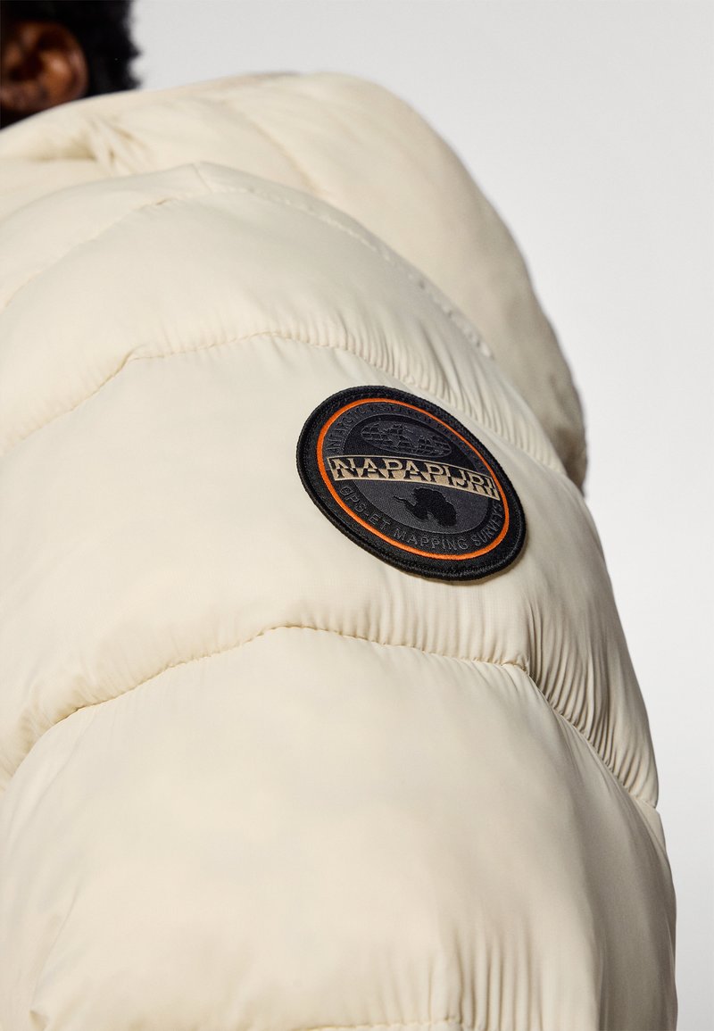 Kurtka puchowa w kremowym kolorze, z pikowanym wzorem. Zawiera okrągłą czarną łatę z pomarańczowymi akcentami i napisem. Miękka, połyskująca tekstura.