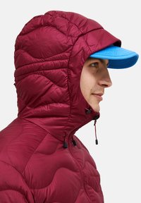 Bordeaux-padded takki, jossa on huppu, ominaisuuksina tikattu pinta ja kiristysnauhat; täydentää sininen lippis, jossa on tasainen lippa.