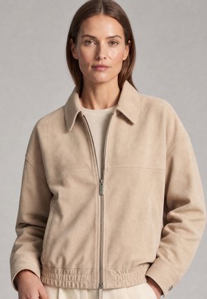 Vrouw met stijlbruin haar die een beige jack met rits draagt over een lichte trui tegen een eentonige grijze achtergrond.