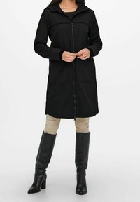 Mujer vestida con un abrigo negro con cremallera y capucha, de pie con confianza en botas negras hasta la rodilla y pantalones beiges frente a un fondo sencillo.