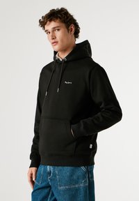 Μαύρη hoodie με κουκούλα και κορδόνια, μπροστινές τσέπες, ριμπ μανσέτες και μικρό λογότυπο στο στήθος, συνδυασμένη με μπλε τζιν παντελόνι.
