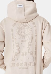 Felpa beige con cappuccio che presenta disegni in rilievo di sole, yin-yang, serpente e testo sul retro. Tessuto morbido con vestibilità rilassata.