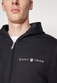 Felpa nera con zip realizzata in tessuto morbido, con il logo "GANT 1949" in bianco e rosso, dotata di cappuccio e zip in metallo lucido.