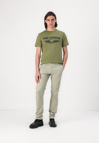 Olivgrünes T-Shirt mit schwarzem "PME LEGEND"-Grafik, kombiniert mit hellbeigen Hosen und schwarzen Schuhen, vor einem weißen Hintergrund.