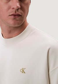 Bílá mikina s žebrovaným kulatým výstřihem. Na levé straně hrudi je malý zlatý vyšívaný znak "cK". Měkká, hladká textury látky.