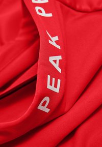 Tessuto atletico rosso con una texture liscia, caratterizzato da un logo bianco con la scritta "PEAK" lungo il bordo di una fascia. Prospettiva ravvicinata.