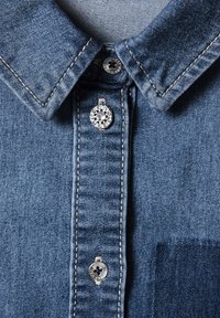 Gros plan sur le col d'une chemise en denim bleu avec trois boutons argentés, dont un décoré de petits strass transparents.