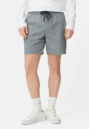 Mann trägt hellgraue Shorts mit schwarzem Kordelzug, weiße Sportsocken, weiße Sneakers und ein langärmliges weißes Hemd, steht vor weißem Hintergrund.