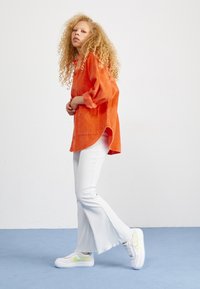 Pomarańczowa koszula z kordurowej tkaniny o luźnym kroju i z zapięciem na guziki, zestawiona z białymi, prążkowanymi spodniami w stylu dzwonów i białymi trampkami z neonowymi akcentami.