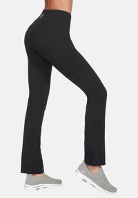 Schwarze Sporthose mit hohem Bund, seitlichen Nahtdetails und Gesäßtaschen, kombiniert mit grauen Slippern.