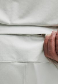 Tela blanca con textura de cuadrícula, que presenta un bolsillo con cremallera. Una mano agarra la tela cerca de la cremallera, mostrando un diseño limpio y un acabado suave.