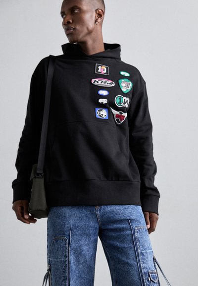 Μαύρη κουκούλα με τσέπη καγκουρό, διακοσμημένη με διάφορες πολύχρωμες ραφές. Φορεμένη με μπλε cargo παντελόνια, που παρουσιάζουν άνετη εφαρμογή και λεπτομέρειες με φερμουάρ.