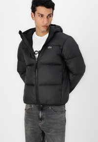 Veste noire matelassée avec capuche, dotée d'une fermeture éclair à l'avant et d'un petit logo de crocodile vert. Fabriquée à partir d'un matériau lisse et léger.