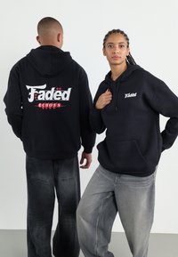 Svart hoodie med framficka, prydd med vitt och rött logotyp på bröstet och på ryggen. Matchas med löst sittande, blekta grå jeans.