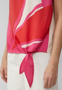 Kleurvolle top met een mengeling van rood en roze en artistieke patronen. De zoom is geknoopt, gemaakt van een soepele, lichte stof.