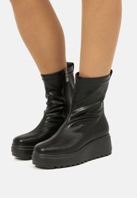 Botas de tobillo de cuero negro con cierre de cremallera, que presentan un diseño elegante, punta redondeada y una suela de cuña con un patrón de goma texturizada.