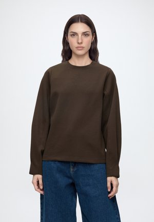 Femme aux longs cheveux bruns portant un t-shirt marron foncé à manches longues et un jean bleu, debout devant un fond blanc uni.