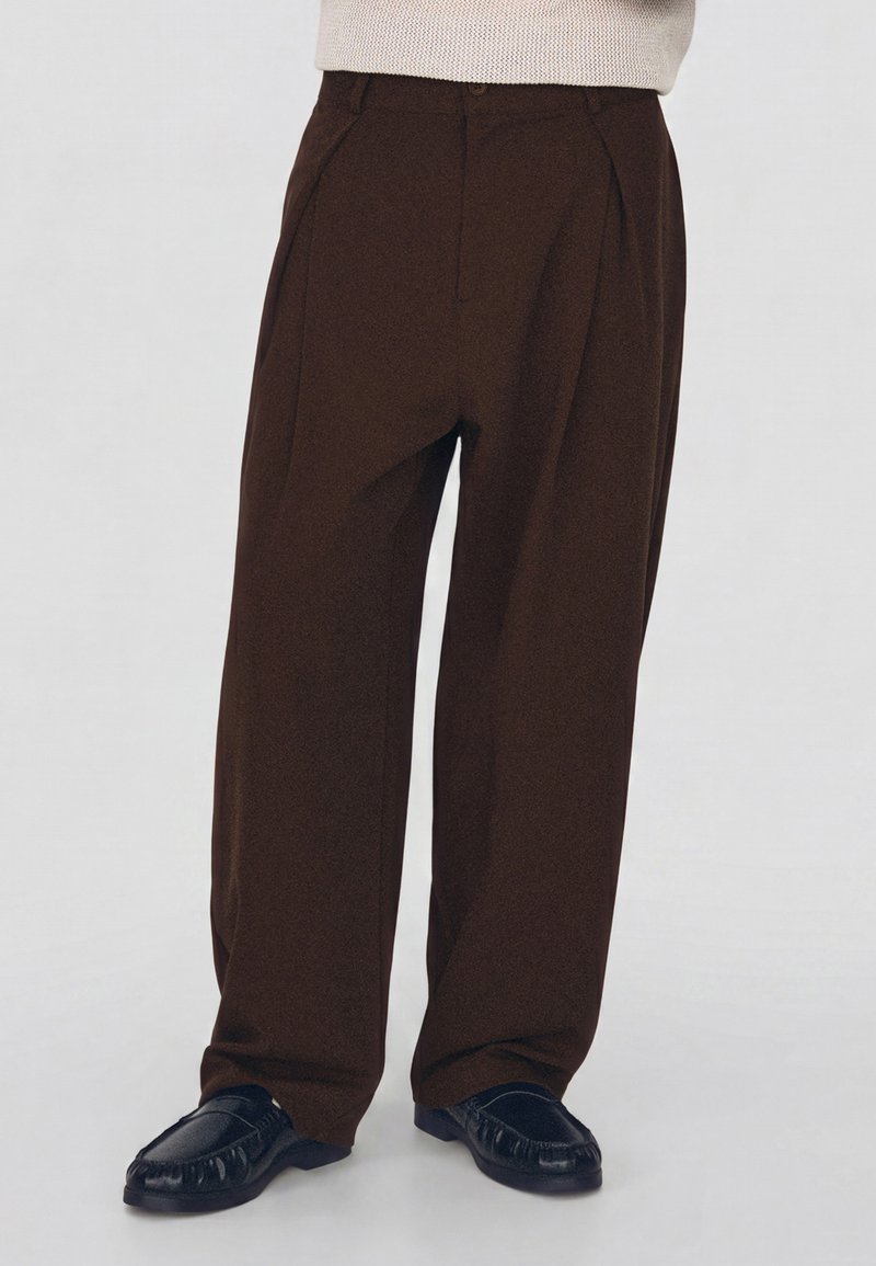 Personne portant un pantalon large marron foncé et des chaussures noires à enfiler, debout devant un fond clair uni.
