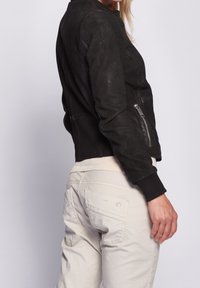 Svart perforerad läderbomberjacka med ribbade ärmslut och midja; har en sidoblixtlås och slät textur. Bärs över ljusa byxor.