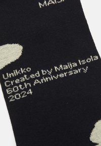 Fekete zoknik nagy fehér virágmintával, "Unikko" felirattal és "Készítette: Maija Isola 80. évforduló 2024" világos szöveggel.