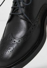 Scarpa da uomo in pelle nera con design brogue, caratterizzata da intricate perforazioni e una finitura liscia. Stile allacciato con punta rotonda.