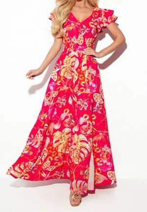 Femme portant une longue robe rose fluide avec des imprimés floraux tropicaux jaunes et orange et des manches flottantes, debout avec une main sur la hanche.