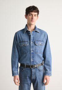 Chemise en denim bleu moyen avec deux poches avant, col boutonné et boutons blancs ; associée à un jean bleu et une ceinture noire.