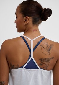 Marinblå och vit sporttopp med racerback-design, texturerat tyg och justerbara band. Inkluderar synliga tatueringar på ryggen.