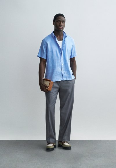 Άνδρας που φοράει ανοιχτό μπλε πουκάμισο με κοντά μανίκια, γκρι παντελόνια και παπούτσια loafers, κρατώντας ένα μικρό καφέ βιβλίο μπροστά σε απλό γκρι φόντο.