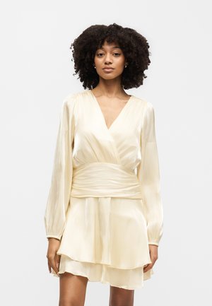 Vrouw met krullend haar die een crèmekleurige satijnen wikkeljurk met lange mouwen en een gelaagde rok draagt, staand tegen een effen witte achtergrond.
