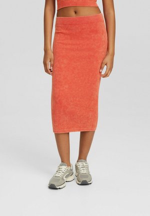 Vrouw draagt een koraalkleurige geribbelde midi rok en een bijpassende cropped top met beige en witte sneakers, staand tegen een effen lichte achtergrond.