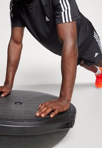 Černé sportovní tričko a šortky s bílými pruhy; ruce se drží černé stabilizační koule; v pozadí jsou viditelné jasně oranžové boty.