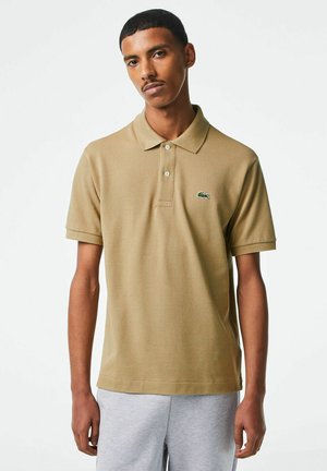 Giovane uomo che indossa una polo a maniche corte color sabbia con un piccolo logo a coccodrillo e pantaloni della tuta grigio chiaro, in piedi contro uno sfondo bianco.