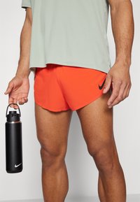 Svart rustfritt stål vannflaske i hånden, iført en lysegrønn kortermet skjorte og klare oransje shorts med logo.