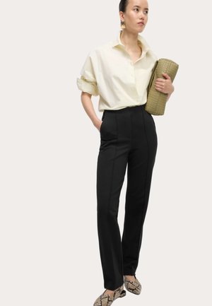 Kvinde iført off-white skjorte med knapper, sorte skræddersyede bukser, mønstrede loafers, holder stor flettet olivengrøn clutch under armen.