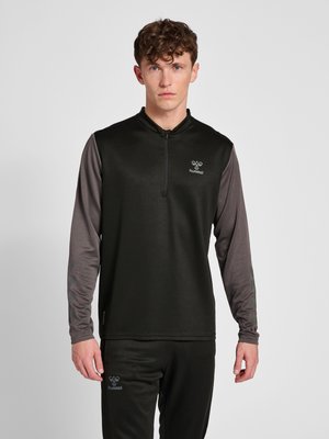 Ung mand iført sort og grå sportsbluse med lynlås og matchende sorte bukser mod ensfarvet baggrund.