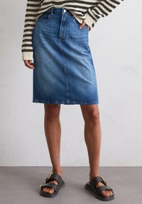 Denim rok met een rechte snit, donkerblauwe kleur, subtiele textuur en een voornaad. Gepaard met zwarte sandalen voorzien van een gouden gesp.