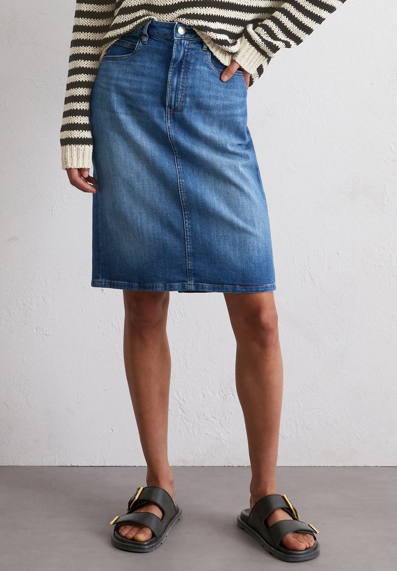 Denim rok met een rechte snit, donkerblauwe kleur, subtiele textuur en een voornaad. Gepaard met zwarte sandalen voorzien van een gouden gesp.