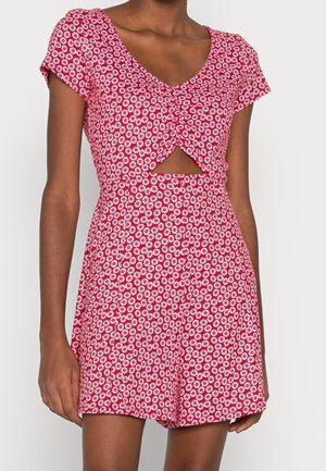 Mujer con un vestido rojo de manga corta con estampado de margaritas blancas y un pequeño recorte triangular debajo del busto, de pie frente a un fondo liso.