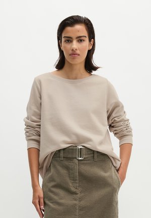 Jeune femme aux cheveux foncés portant un chemisier beige à manches longues et une jupe verte en velours côtelé avec une ceinture, debout avec une main dans la poche.