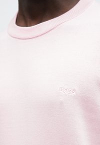 Κοντινό πλάνο ενός ατόμου που φοράει ένα ανοιχτό ροζ πλεκτό πουλόβερ �με στρογγυλή λαιμόκοψη και μικρό κεντημένο λογότυπο "BOSS" στο στήθος.