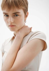 Top de crema ajustado con mangas cortas y cuello; textura suave, brillo sutil; la modelo está sosteniendo su brazo, mostrando un pendiente de aro plateado.