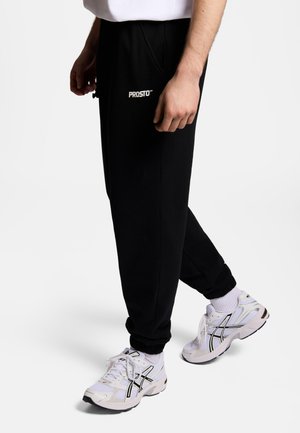 Mężczyzna w czarnych spodniach dresowych z logo "PROSTO" oraz białych sneakersach z czarnymi detalami, stojący na jednolitym białym tle.